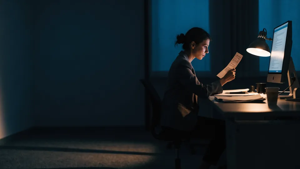 A finance analyst comparing a paper transfer receipt against a monitor at night
