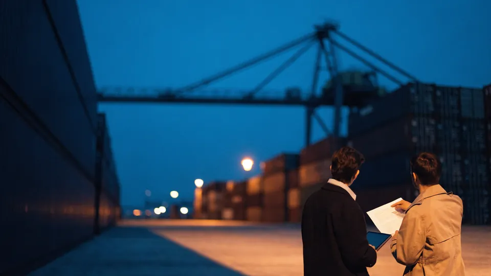 Two professionals reviewing shipping documents at a container terminal at dusk